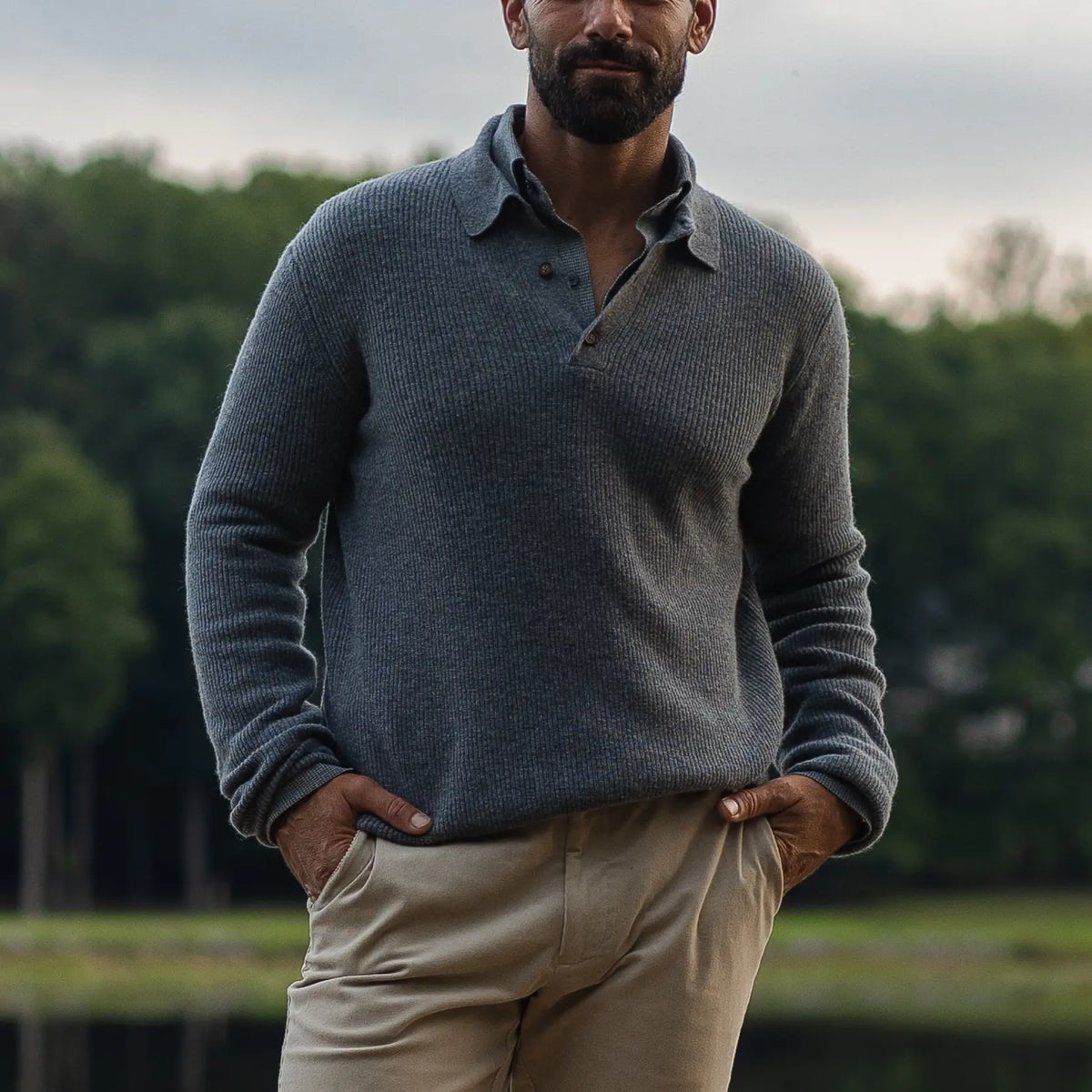 A man wearing a dark gray ribbed knit polo sweater and beige trousers, standing outdoors with his hands in his pockets against a blurred background of trees and water.