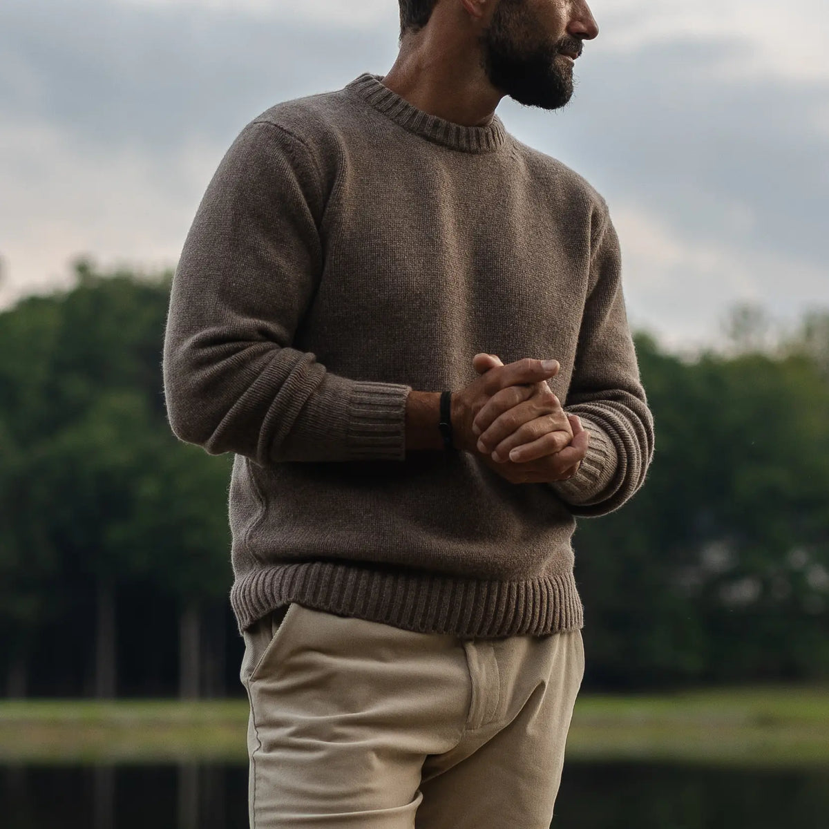 A man wearing a brown knit crewneck Italian merino wool sweater and beige trousers, standing outdoors with a blurred background of trees and water.