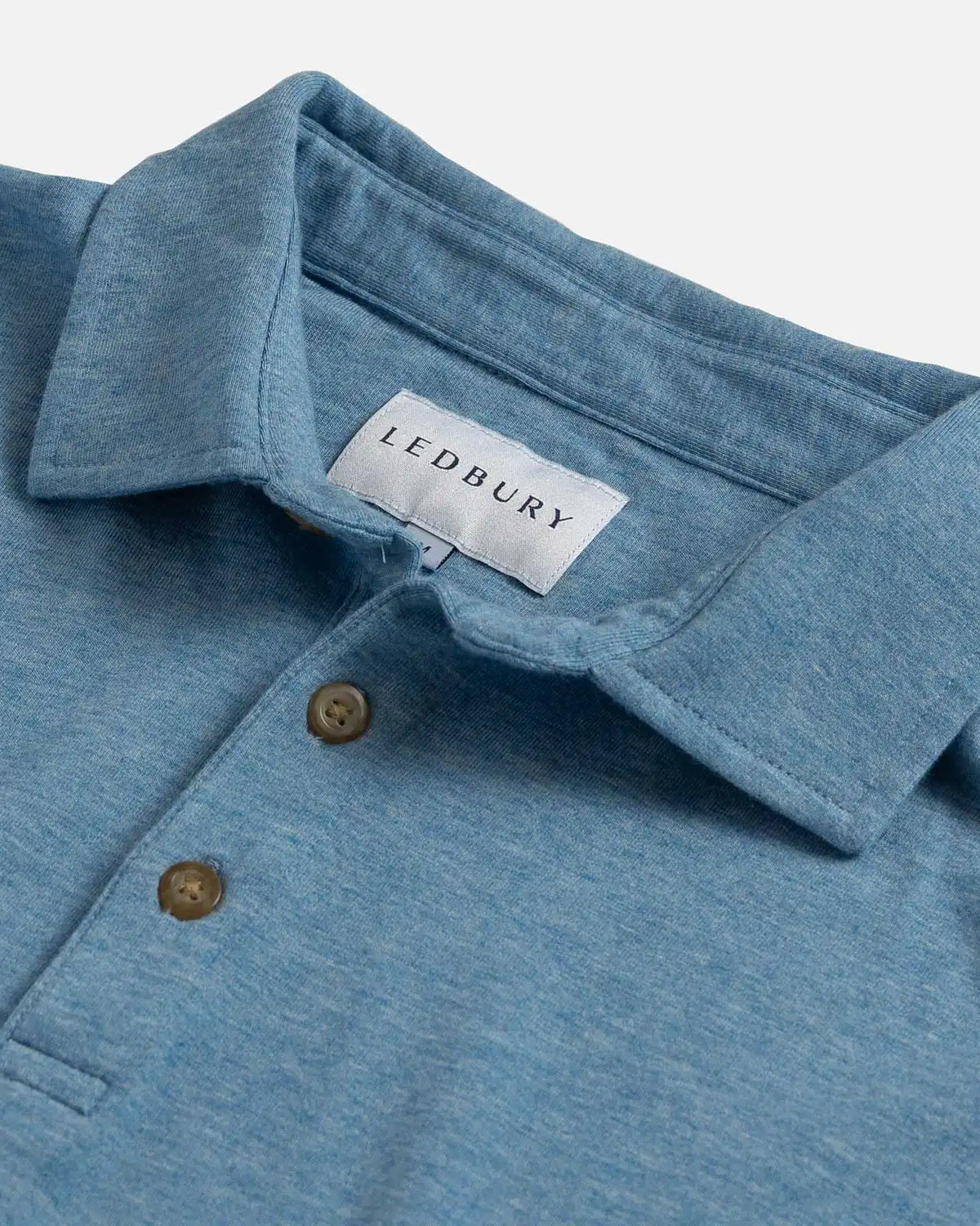 Close-up of a heathered blue polo shirt collar with two brown buttons and a visible LEDBURY neck label on a light gray background.