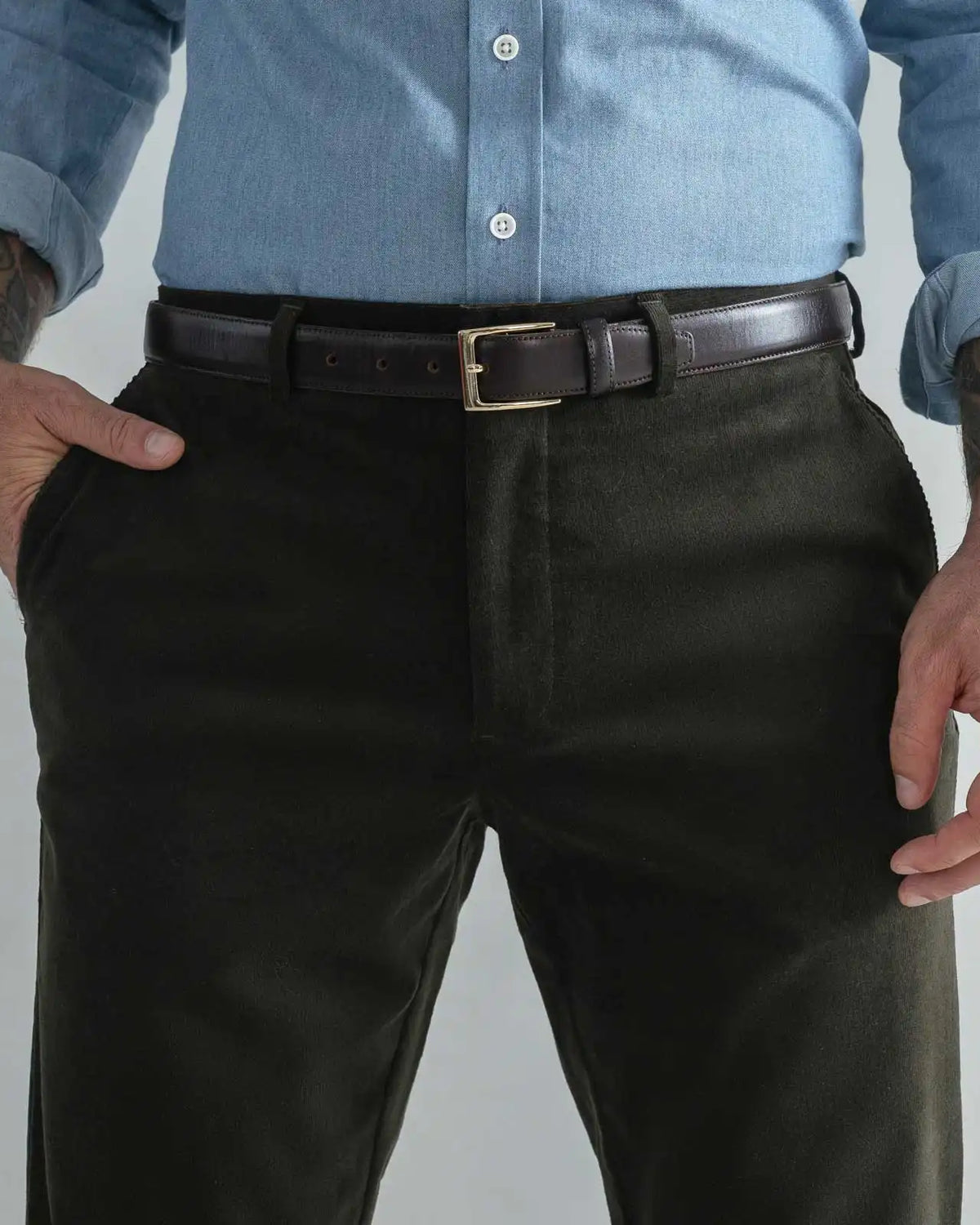 A close-up of a man wearing dark olive green corduroy trousers, a dark brown leather belt with a gold buckle, and a light blue button-down shirt. His hands are in his pockets, and he has visible tattoos on his arms.