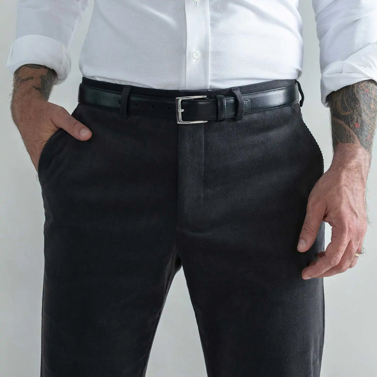 A close-up of a man wearing dark charcoal gray corduroy trousers, a black leather belt with a silver buckle, a white button-down shirt, and tattoos on both arms, set against a plain light gray background.