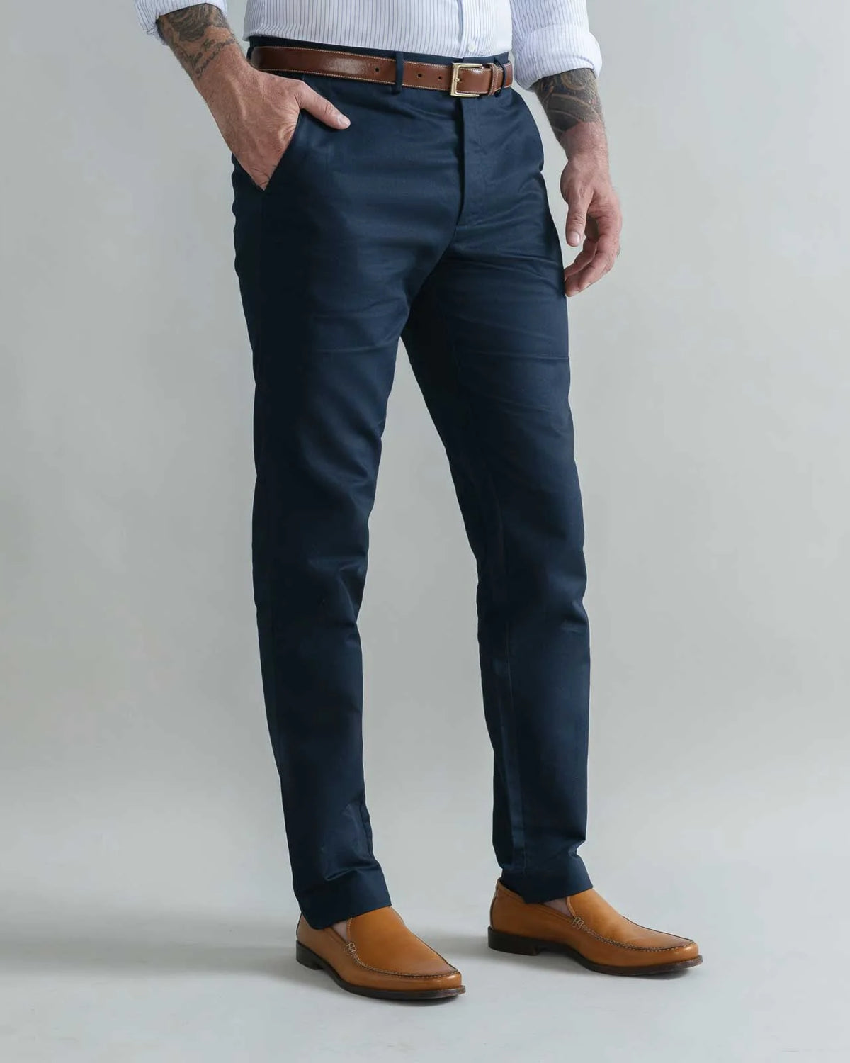 A man wearing slim-fit navy blue trousers, styled with a brown leather belt, a white shirt, and tan leather shoes, standing against a light gray studio background.