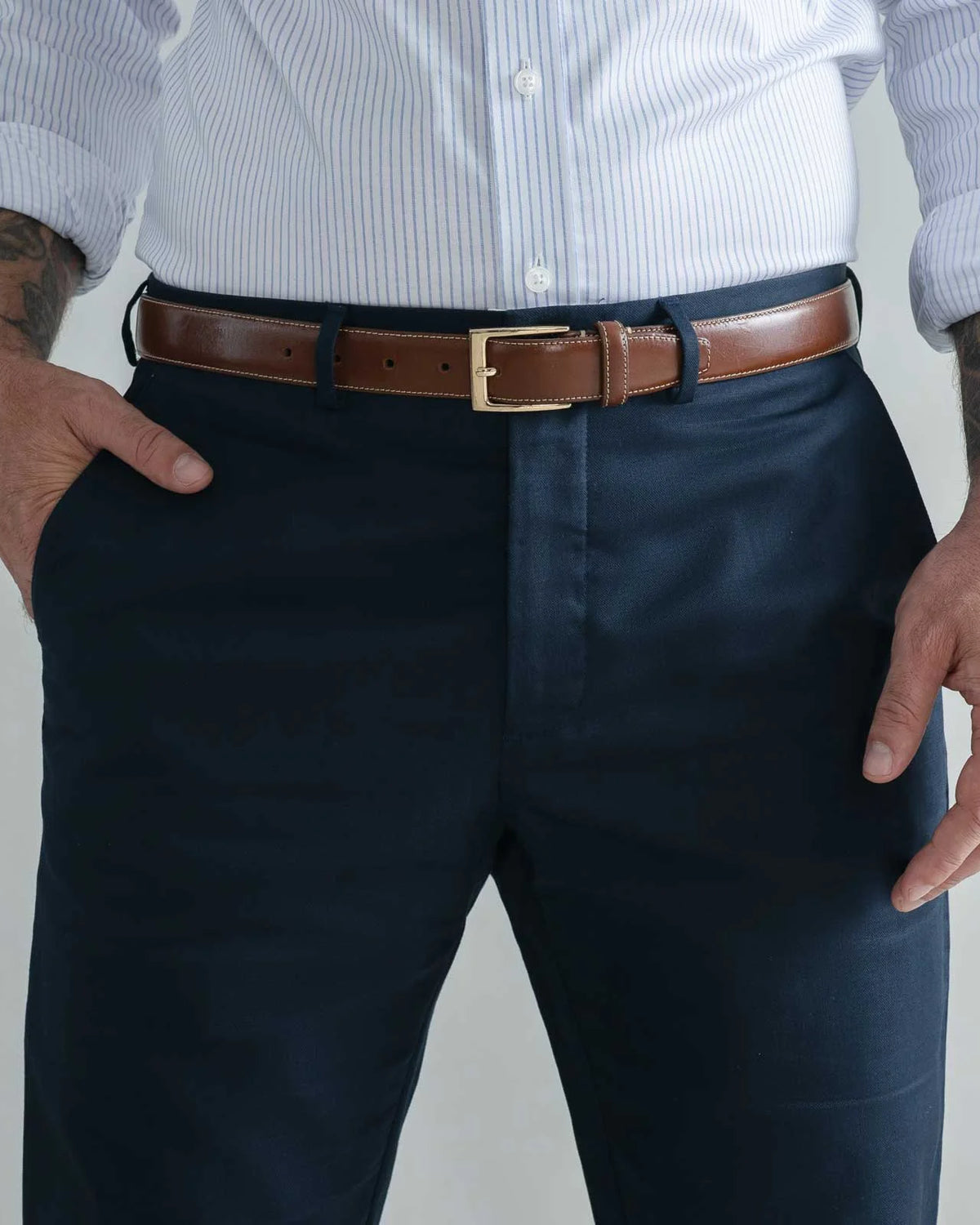 A man wearing navy blue dress pants, a brown leather belt with a gold buckle, and a white and blue striped button-down shirt, shown from the chest down.