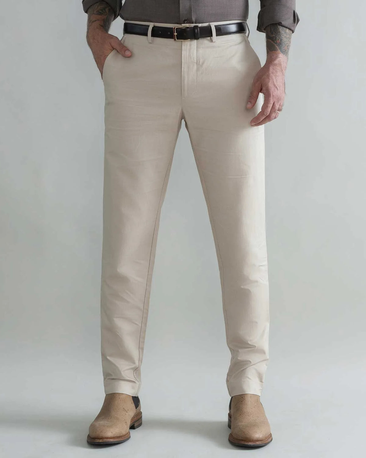 A man wearing khaki slim-fit chinos with a black belt, a brown shirt, and tan boots, standing against a light gray studio background.