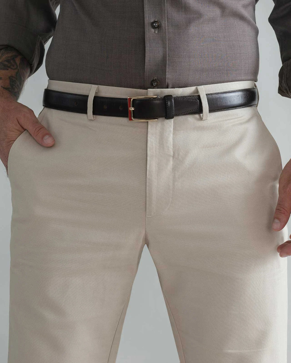 A close-up of a man wearing light khaki chinos with a black leather belt and a dark gray button-down shirt, cropped from the chest down.