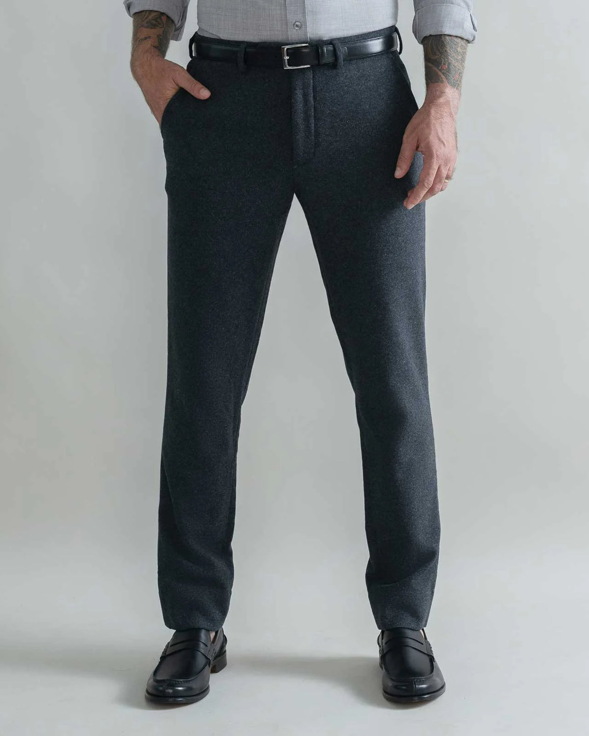 A man wearing dark charcoal gray wool trousers, a black leather belt, a light gray shirt, and black leather loafers, standing against a light gray studio background.