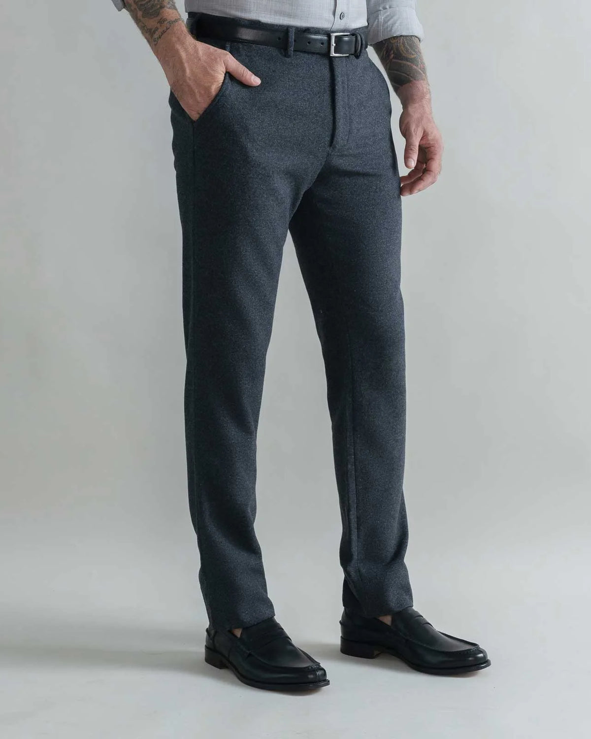 A man wearing dark gray heathered wool trousers, a black belt, a light gray shirt, and black leather loafers, standing against a light gray studio background.