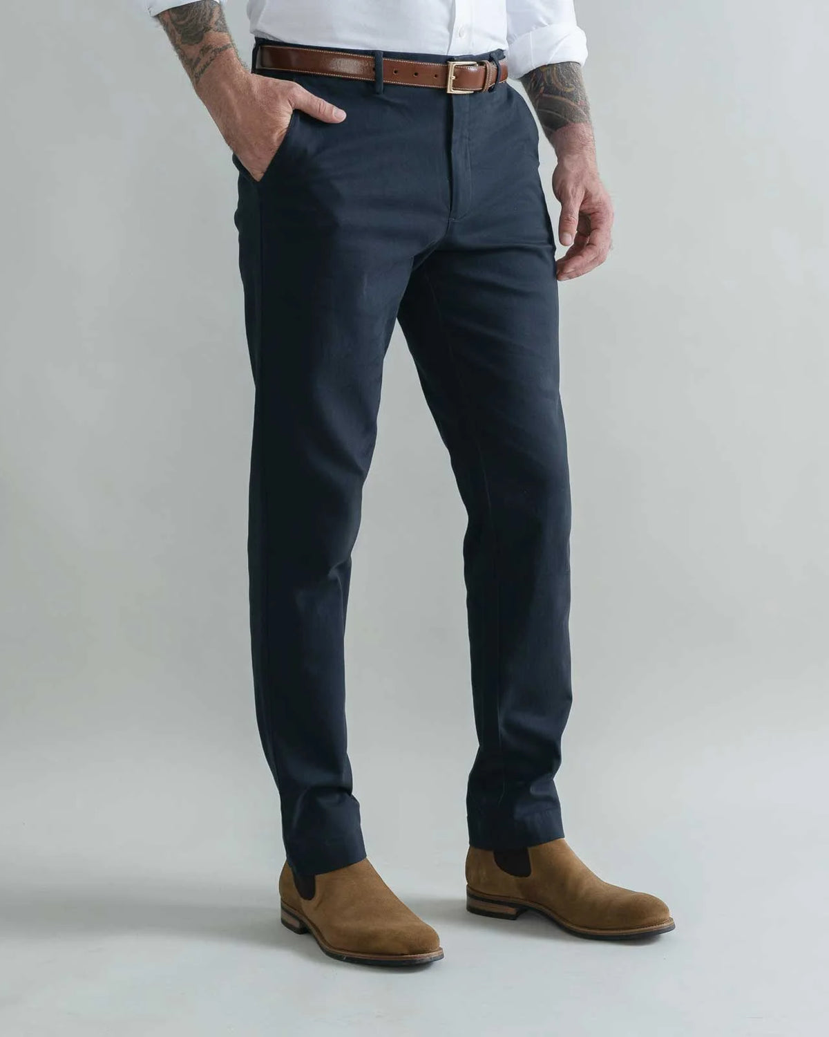 A man wearing navy blue slim-fit trousers, styled with a brown leather belt, a white shirt, and brown suede Chelsea boots, standing against a light gray studio background.