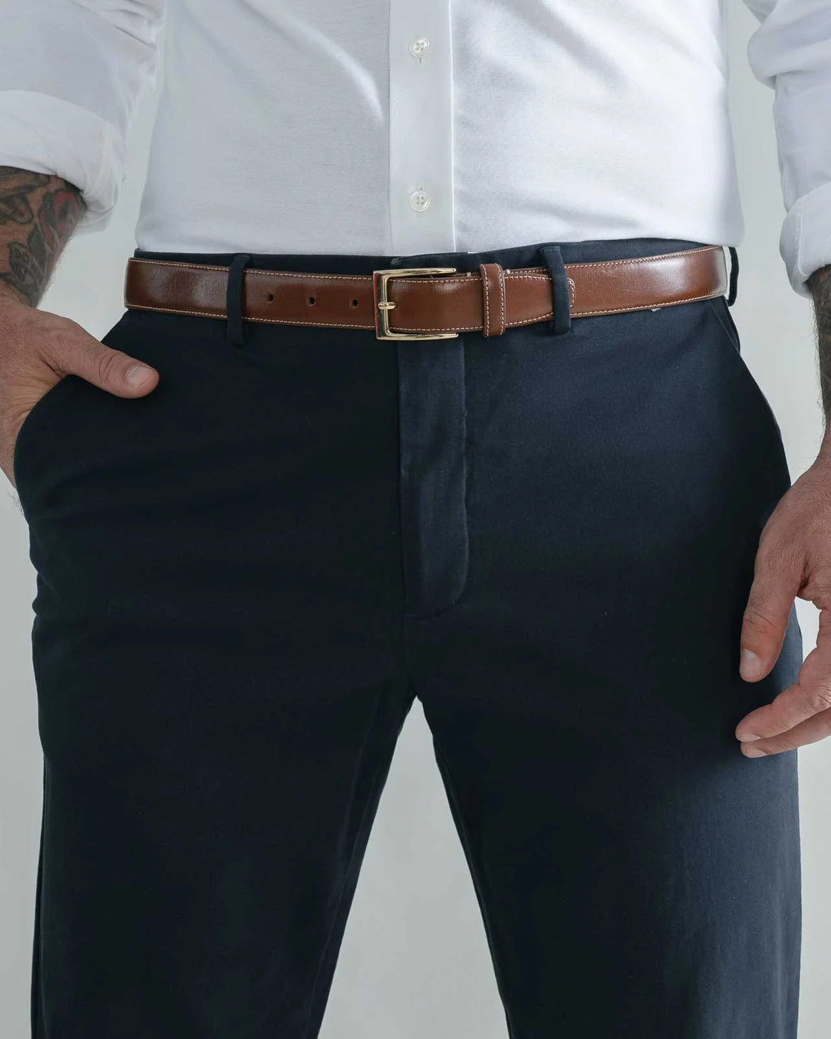 A man wearing dark navy trousers, a brown leather belt with a gold buckle, a white button-down shirt, and tattoos on both arms, standing against a light gray studio background.