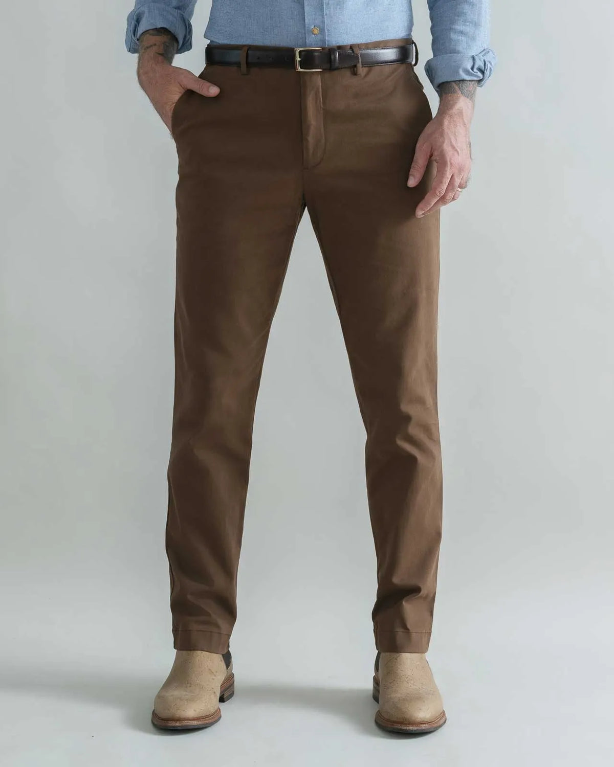 A person wearing slim-fit brown trousers, a black belt, a light blue button-up shirt, and tan boots, standing against a plain light gray studio background.
