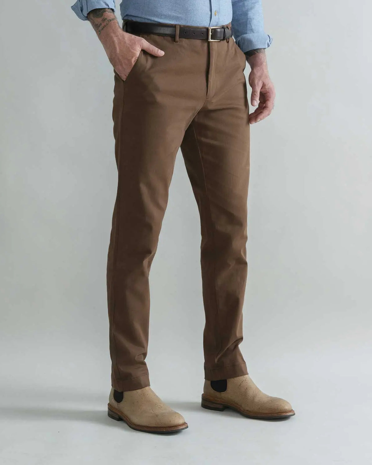 A man wearing slim-fit brown trousers, a dark brown belt, a light blue shirt, and tan suede boots, standing against a light gray studio background.
