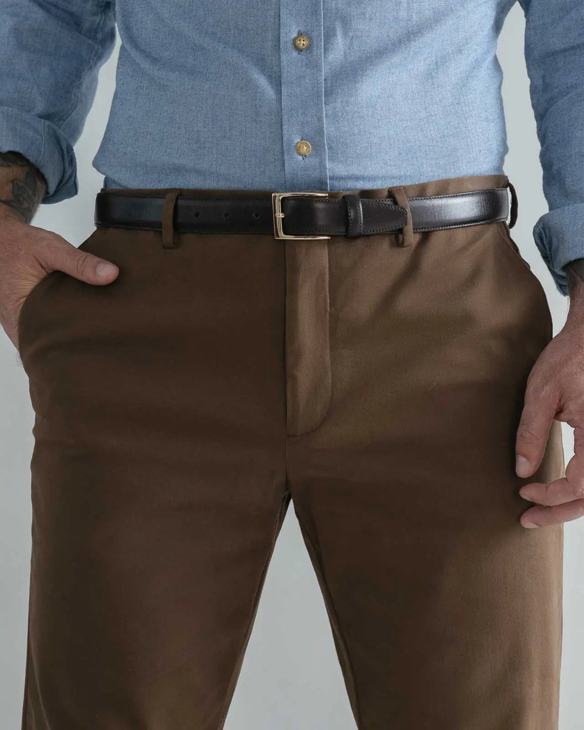 A close-up of a man wearing brown dress pants with a black leather belt and a light blue button-down shirt, shown from the chest down against a plain light gray background.