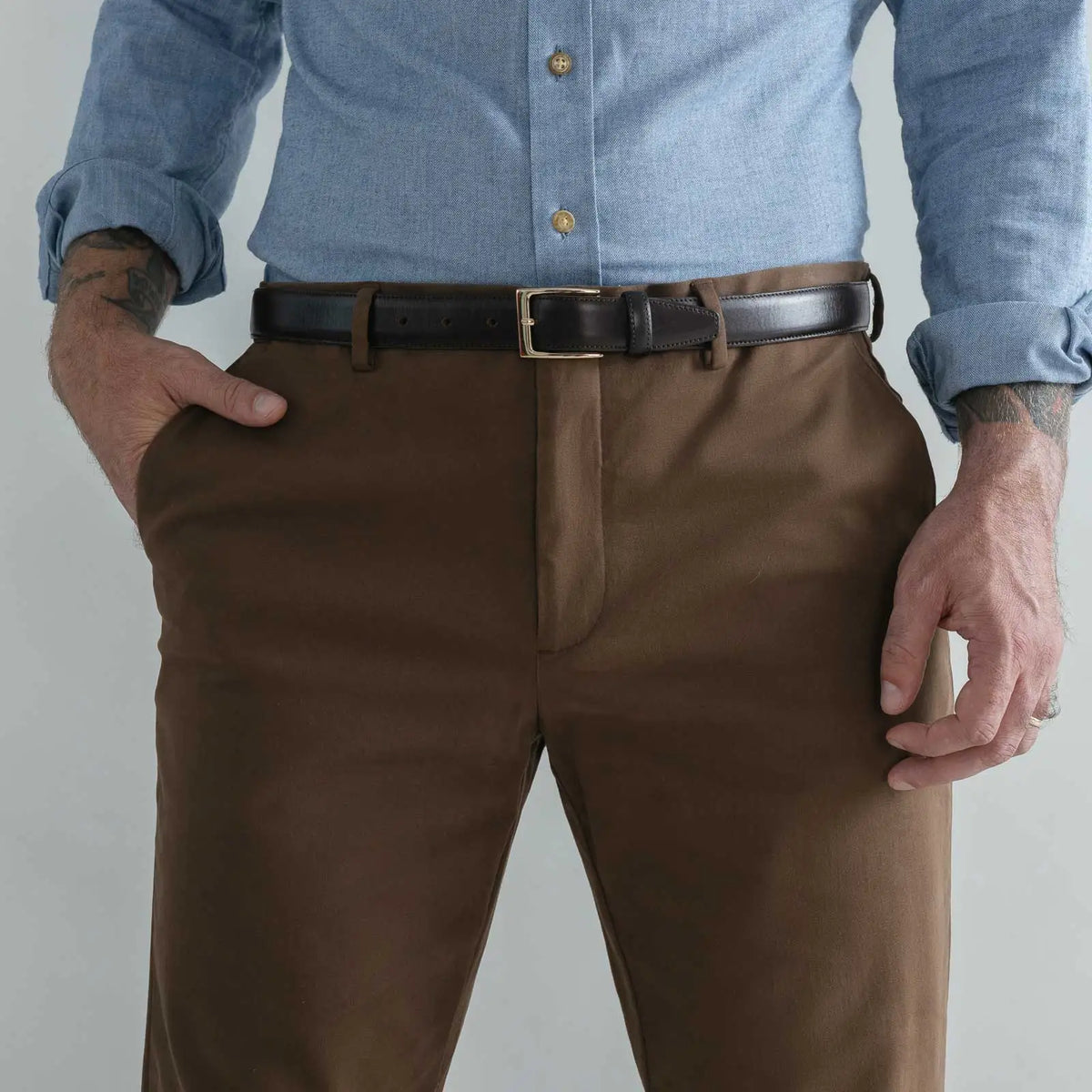 A close-up of a man wearing brown dress pants with a black leather belt and a light blue button-down shirt, shown from the chest down against a plain light gray background.