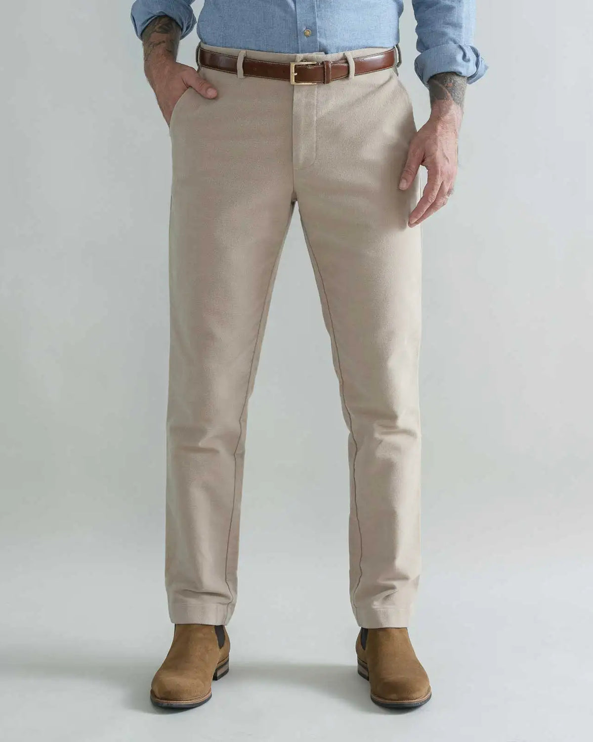 A man wearing tan moleskin chino pants with a brown belt, a light blue button-down shirt, and brown boots, standing against a light gray studio background.