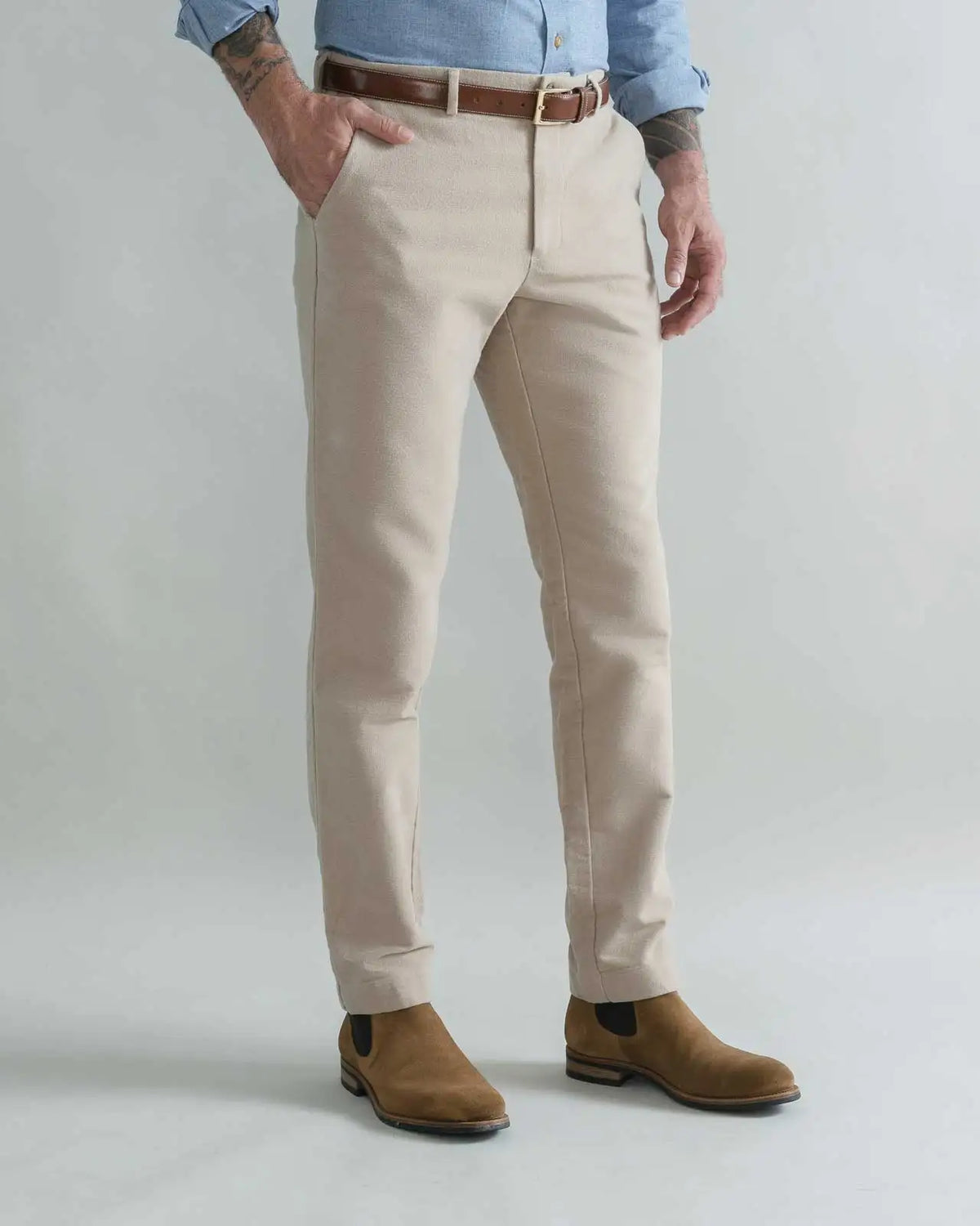 A man wearing tan moleskin chinos with a brown belt, a light blue shirt, and brown suede boots, standing against a light gray studio background.