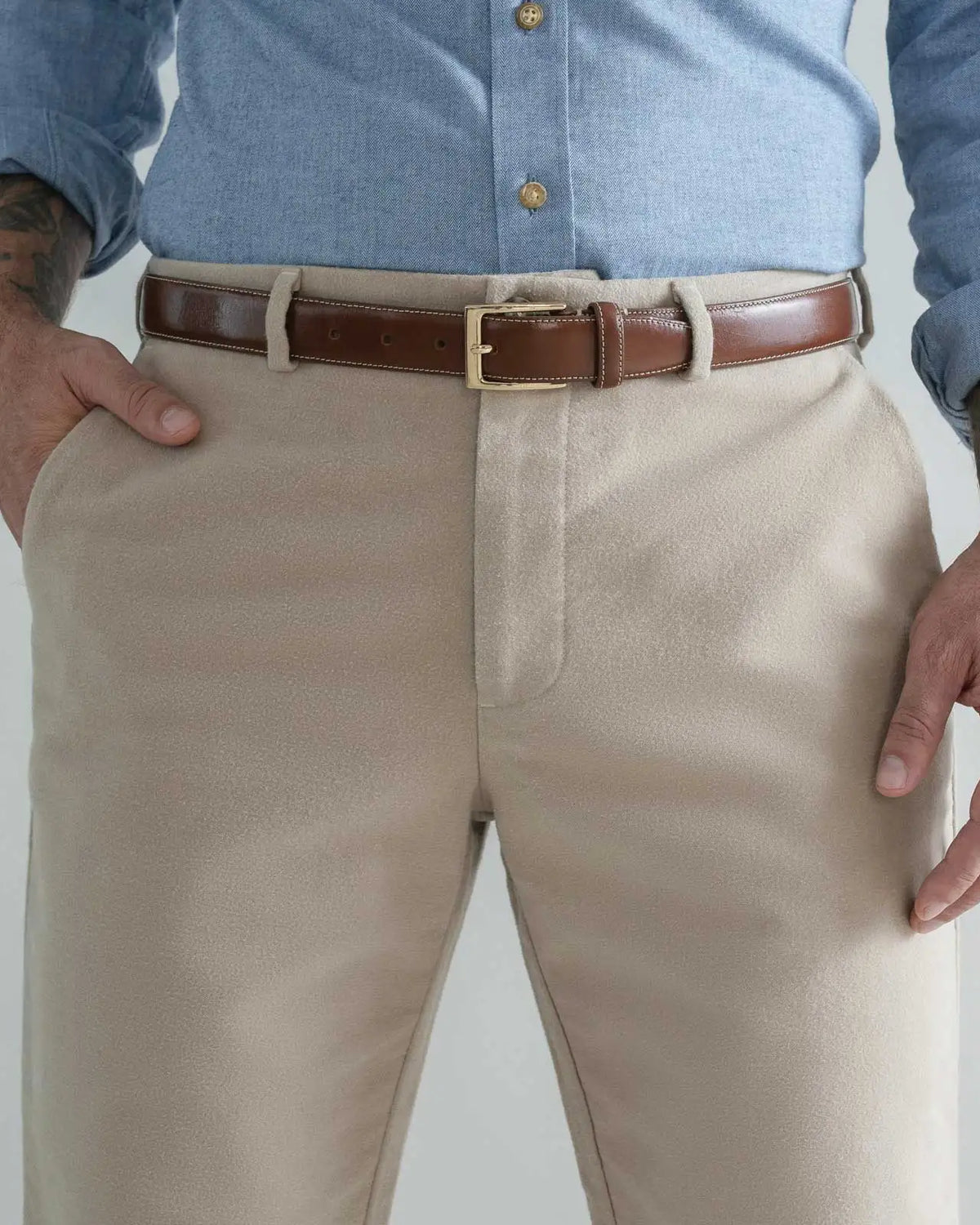A close-up of a man wearing tan moleskin dress pants, a brown leather belt with a gold buckle, and a light blue button-down shirt. His hands are in his pockets, and the background is a clean, light gray studio backdrop.