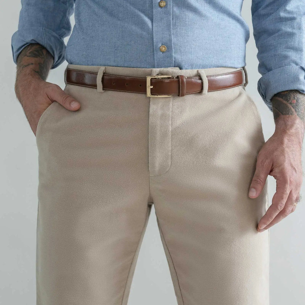 A close-up of a man wearing tan moleskin dress pants, a brown leather belt with a gold buckle, and a light blue button-down shirt. His hands are in his pockets, and the background is a clean, light gray studio backdrop.