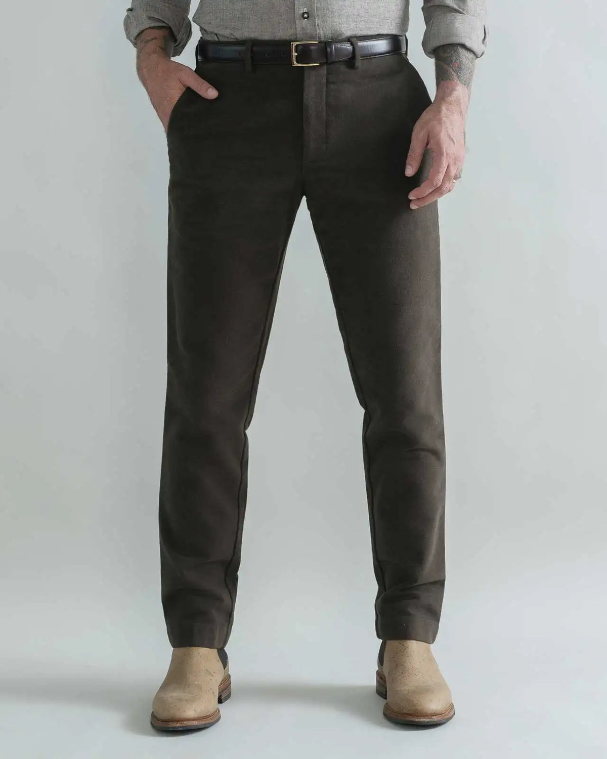 A person wearing slim-fit dark brown moleskin trousers, styled with a black belt, a gray shirt, and beige boots, standing against a light gray studio background