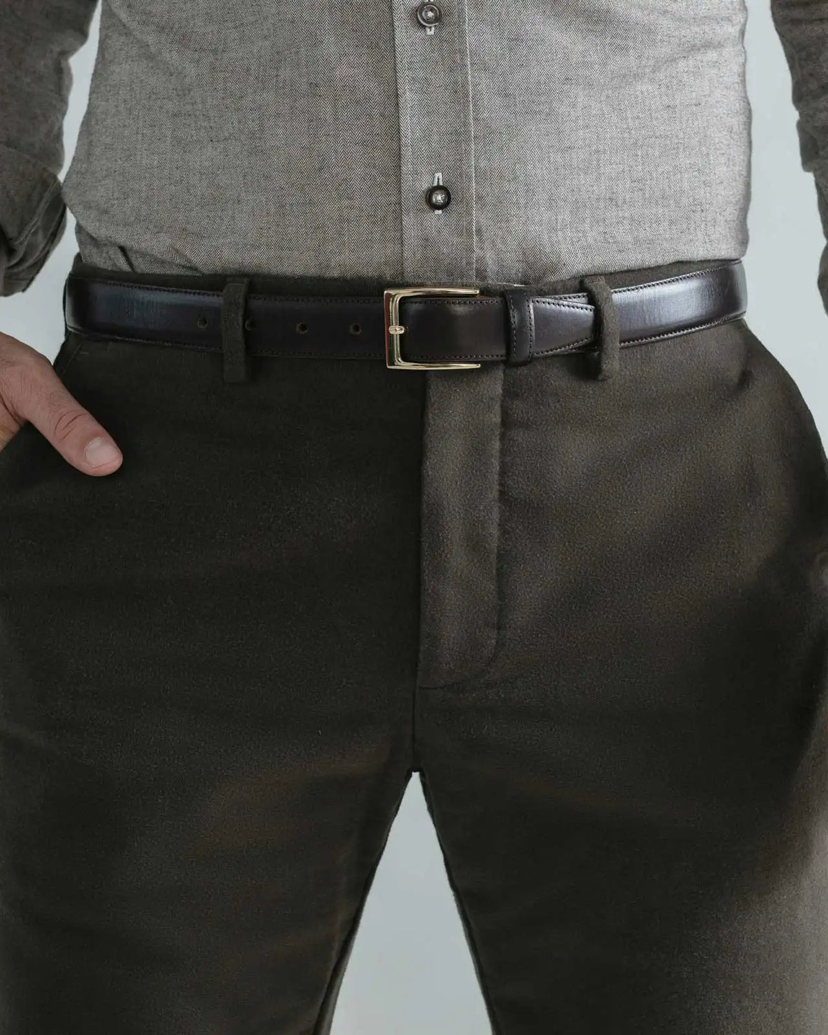 A close-up of a man wearing dark brown moleskin trousers, a black leather belt with a gold buckle, a light gray button-down shirt, and a dark shirt underneath.