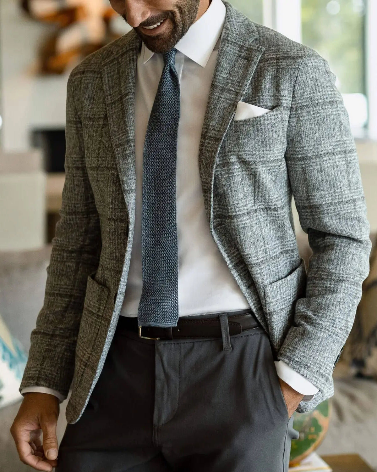 A man wearing a gray and white plaid blazer, white dress shirt, textured blue tie, and dark gray trousers, standing in a modern living room setting.