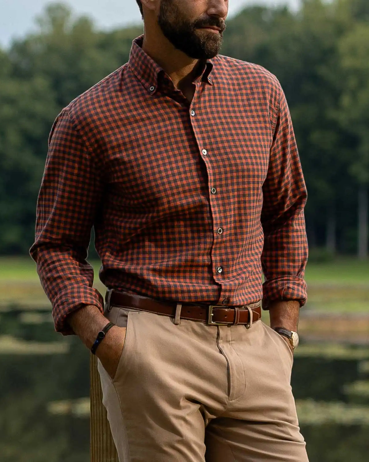 A man wearing a rust-orange and brown checkered long-sleeve button-down shirt, khaki pants, and a brown belt, standing outdoors near a lake with trees in the background.