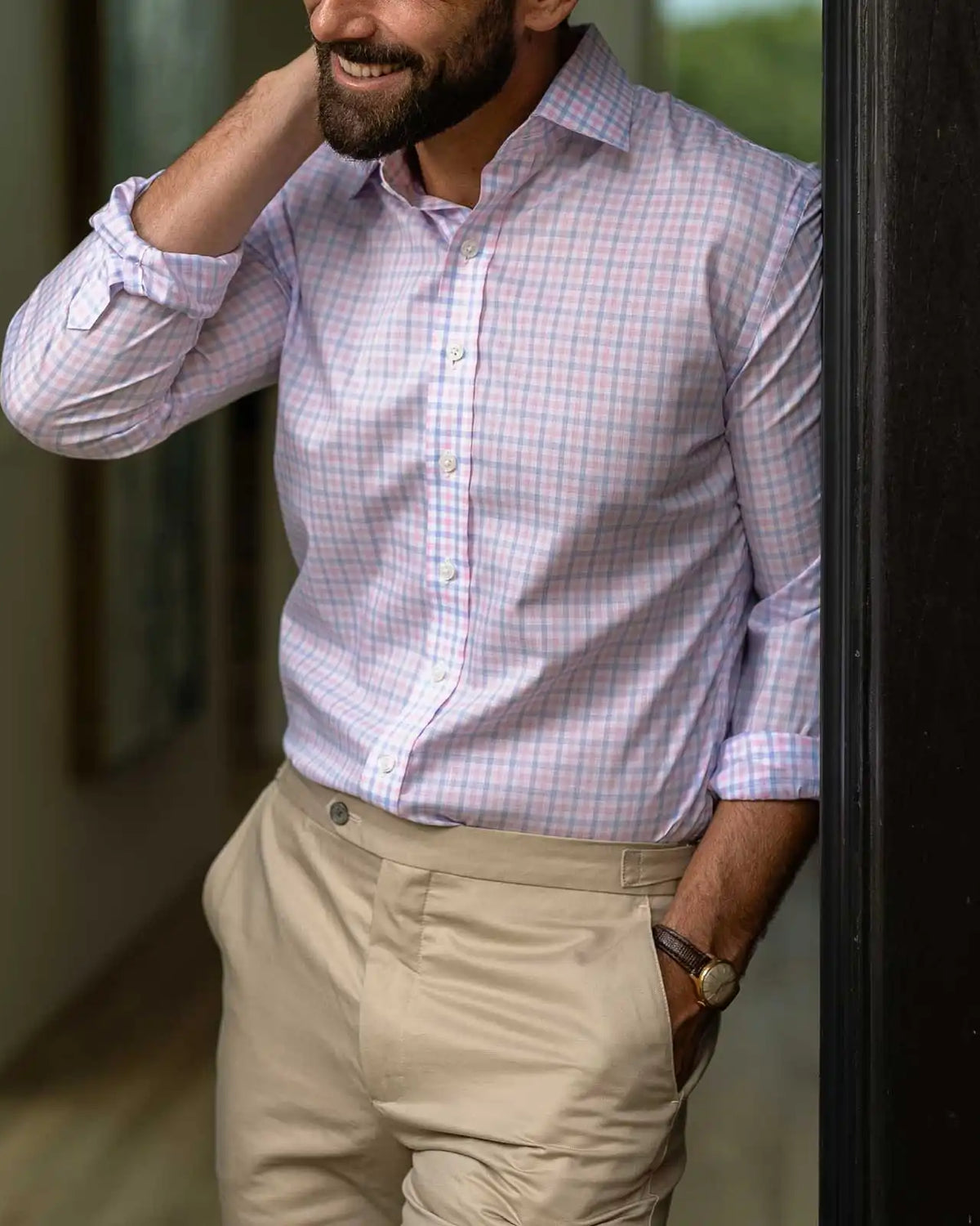 A man wearing a light pink, light blue, and white checkered button-down shirt with beige trousers, standing in a doorway.