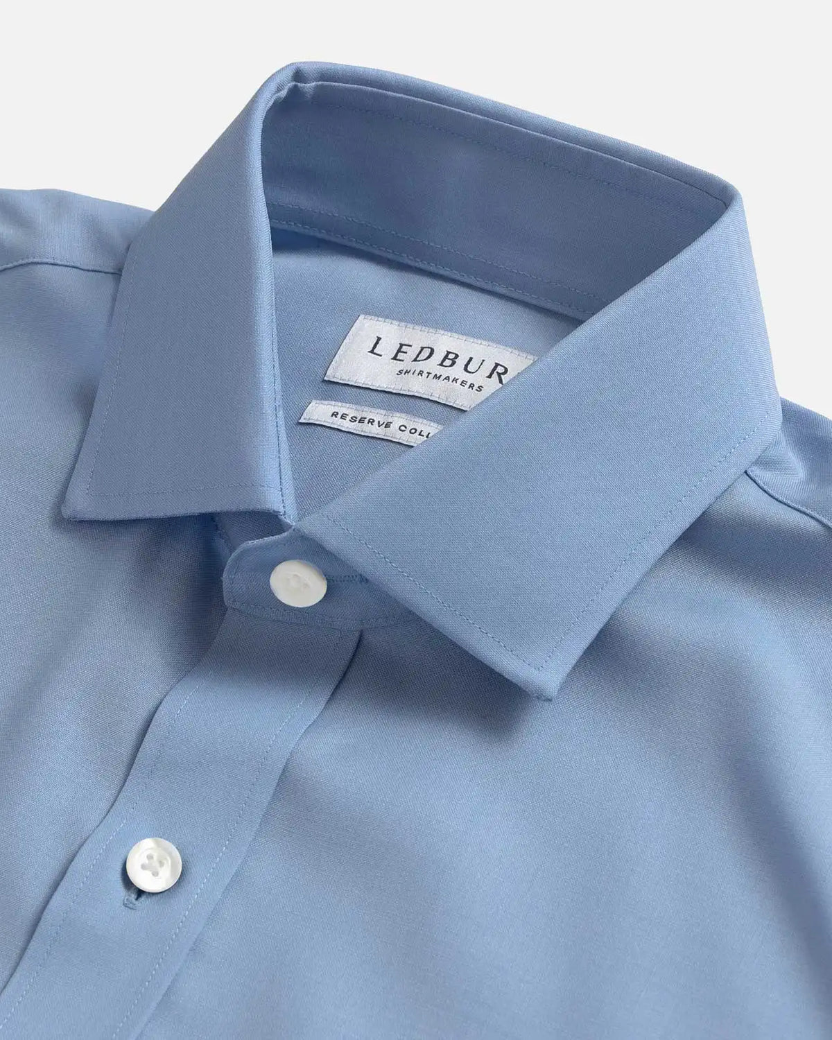 A close-up of a light blue dress shirt collar with a visible LEDBURY brand label, showing the collar point, buttons, and fabric texture against a clean light background.