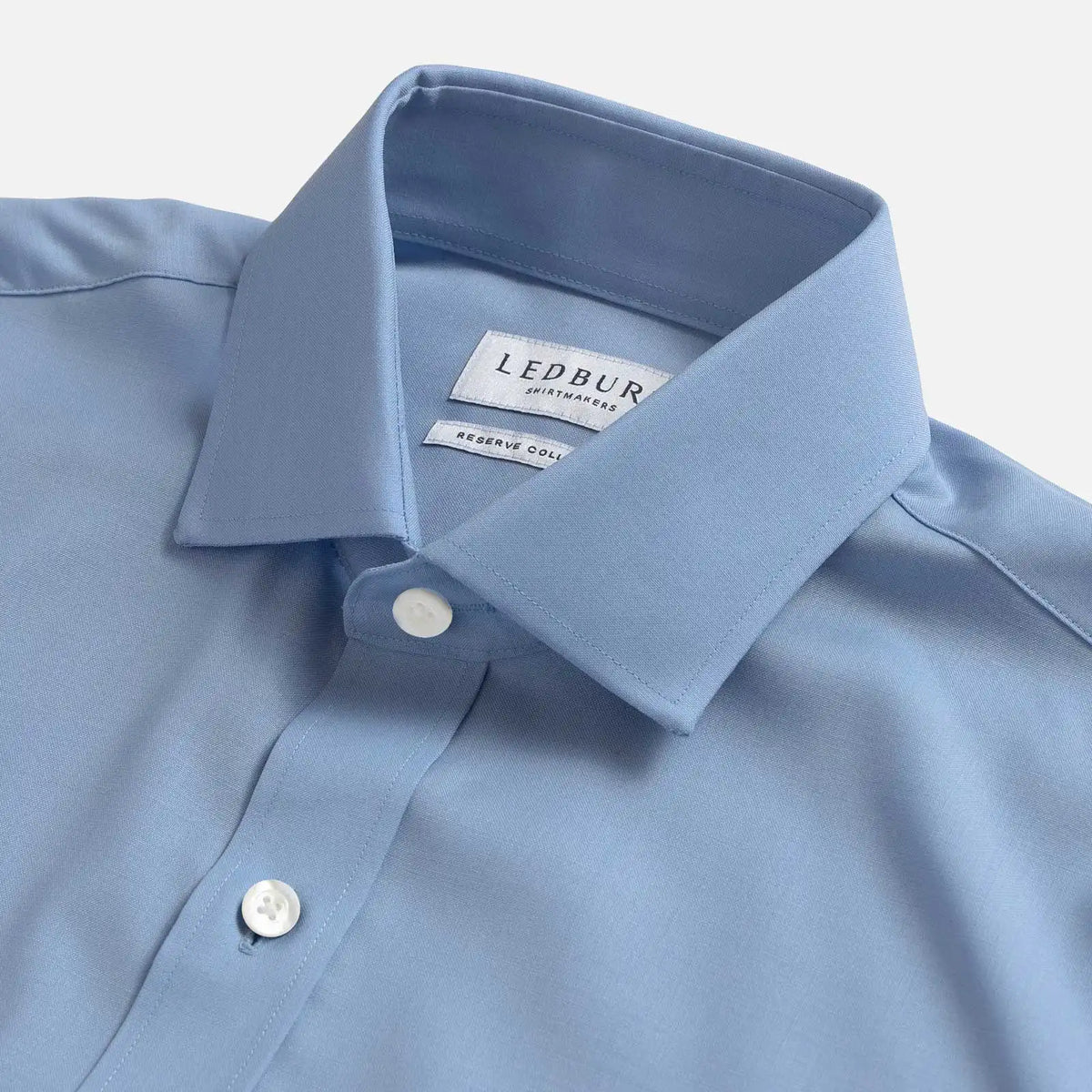 A close-up of a light blue dress shirt collar with a visible LEDBURY brand label, showing the collar point, buttons, and fabric texture against a clean light background.
