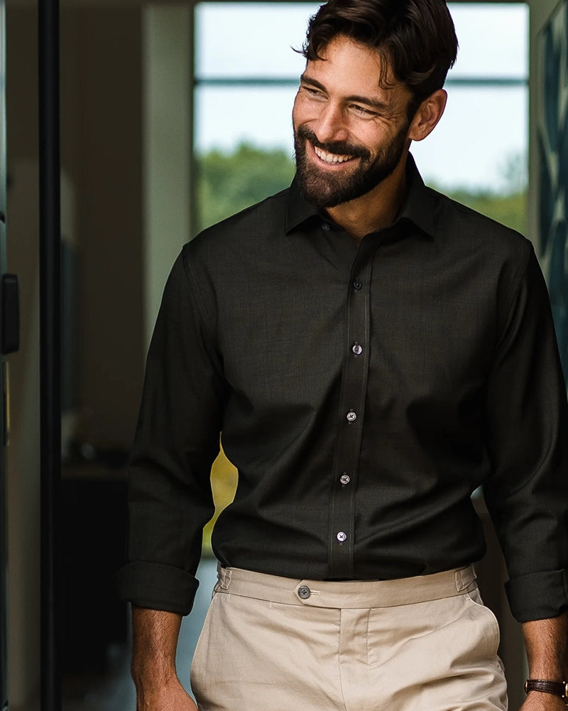 A smiling man wearing a dark green long-sleeve fine Merino button down shirt and beige trousers, standing in a modern indoor setting with a window in the background.