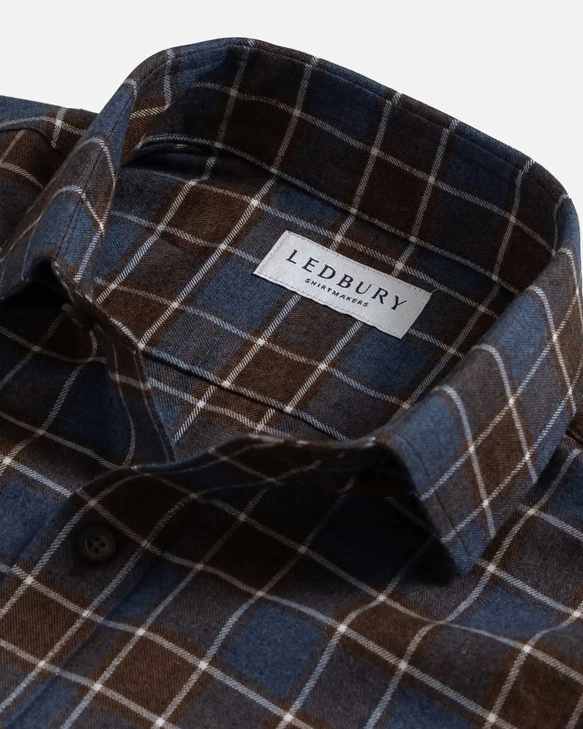 Close-up of a plaid button-down shirt collar with a visible LEDBURY brand label. The fabric is a mix of dark brown, slate blue, and thin white grid lines.