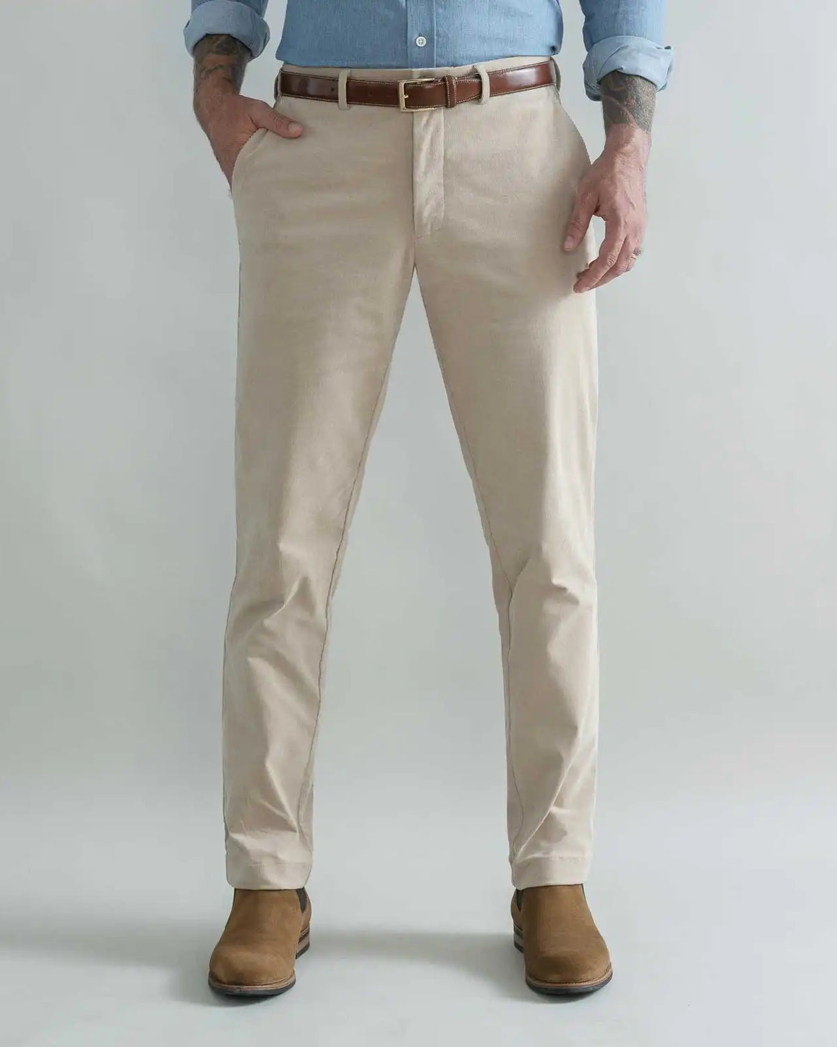 A man wearing tan corduroy chino pants with a brown belt, a light blue button-down shirt, and brown boots, standing against a light gray studio background.
