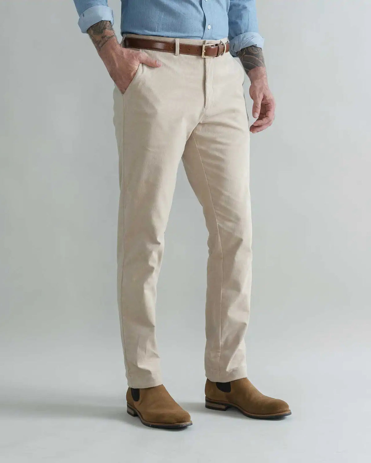 A man wearing tan slim-fit corduroy chinos with a brown belt, a light blue button-down shirt, and brown suede boots, standing against a light gray studio background.