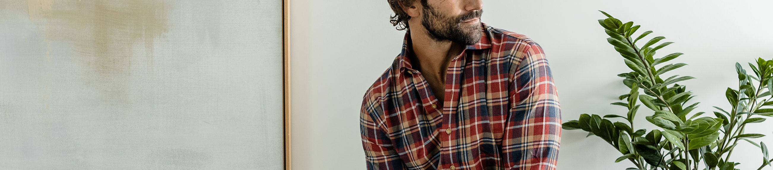 Man wearing a plaid shirt standing next to a plant against a light wall.