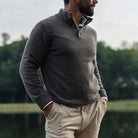 Man wearing a taupe light brown mock neck Italian merino sweater. He is standing outside in front of a lake with a blurred background. 