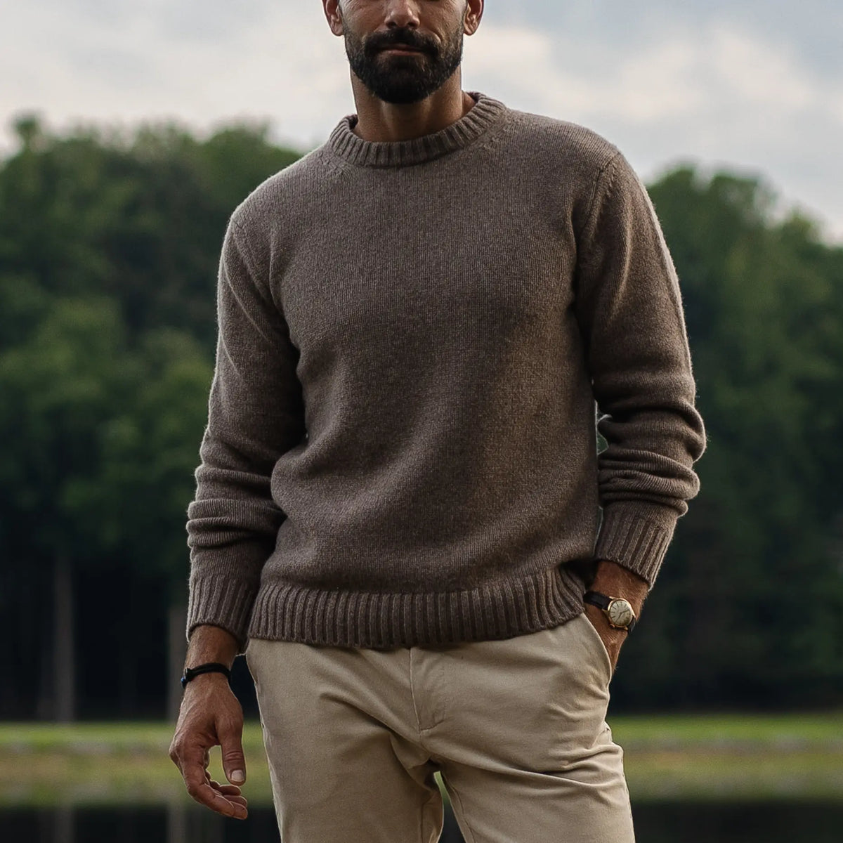 A man wearing a brown knit 100% merino wool crewneck sweater and beige trousers, standing outdoors with a blurred background of trees and water.
