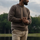 A man wearing a brown knit crewneck Italian merino wool sweater and beige trousers, standing outdoors with a blurred background of trees and water.