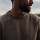 A close-up of a man wearing an Italian merino wool brown knit crewneck sweater, shown from the chest up against a soft, blurred outdoor background.