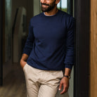 A man wearing a navy blue long-sleeve crewneck sweater and beige trousers, leaning against a dark doorway in an indoor setting.