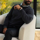 A man wearing a dark charcoal gray knit sweater over a light gray collared shirt, seated in a cream boucle armchair.