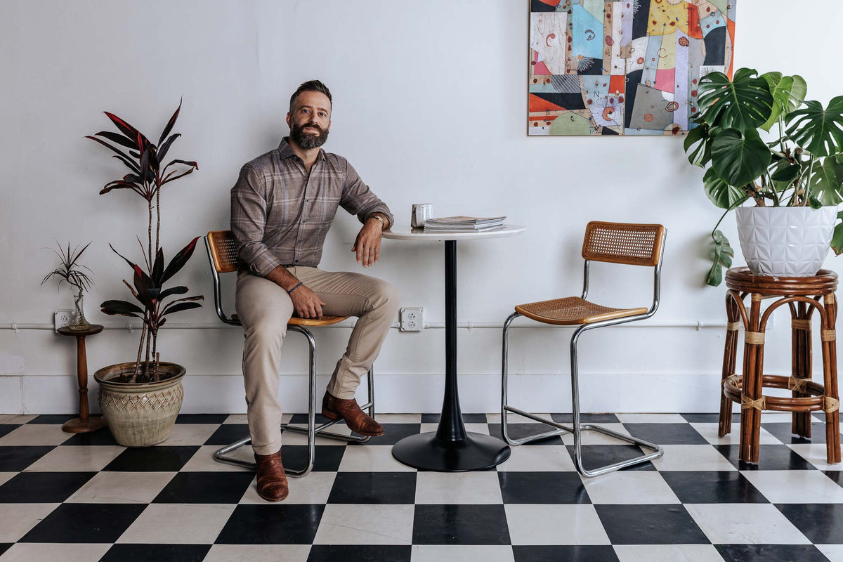 Man sitting at a small table in a room with checkered floor, plants, and colorful wall art.