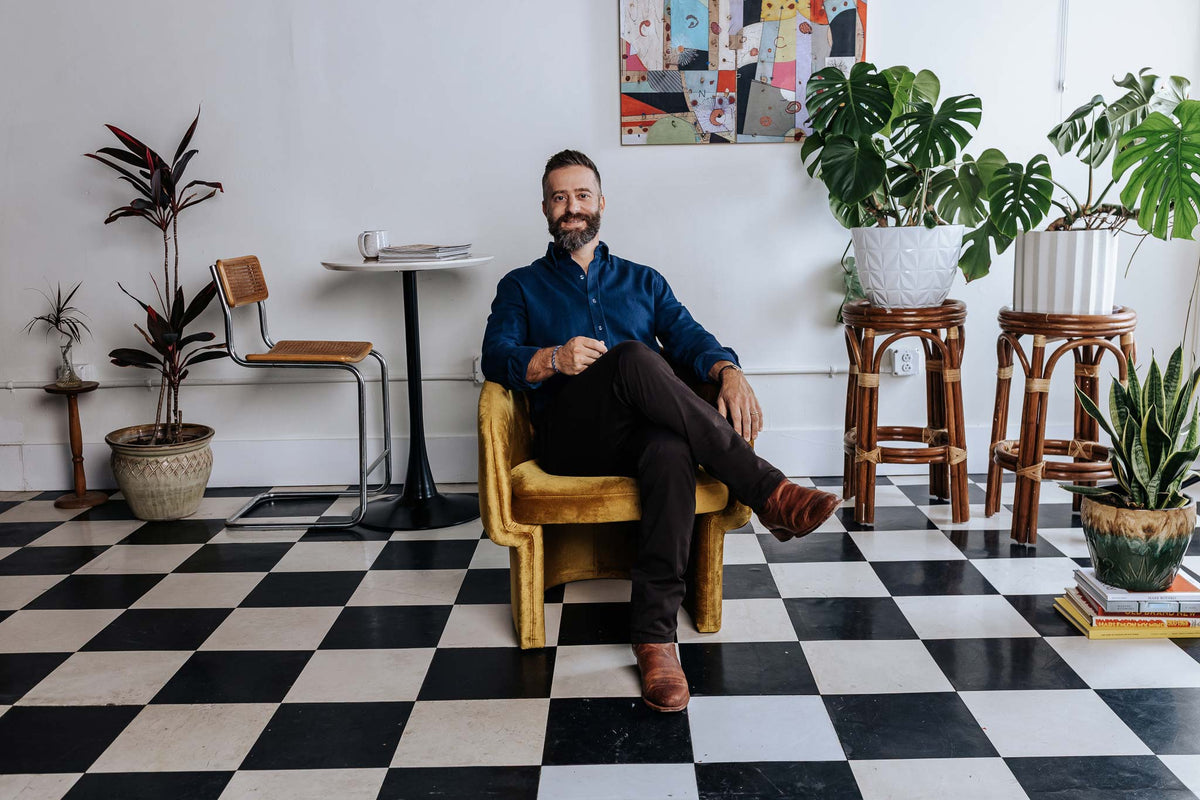 Man sitting in a chair on a black and white checkered floor with plants and furniture around.
