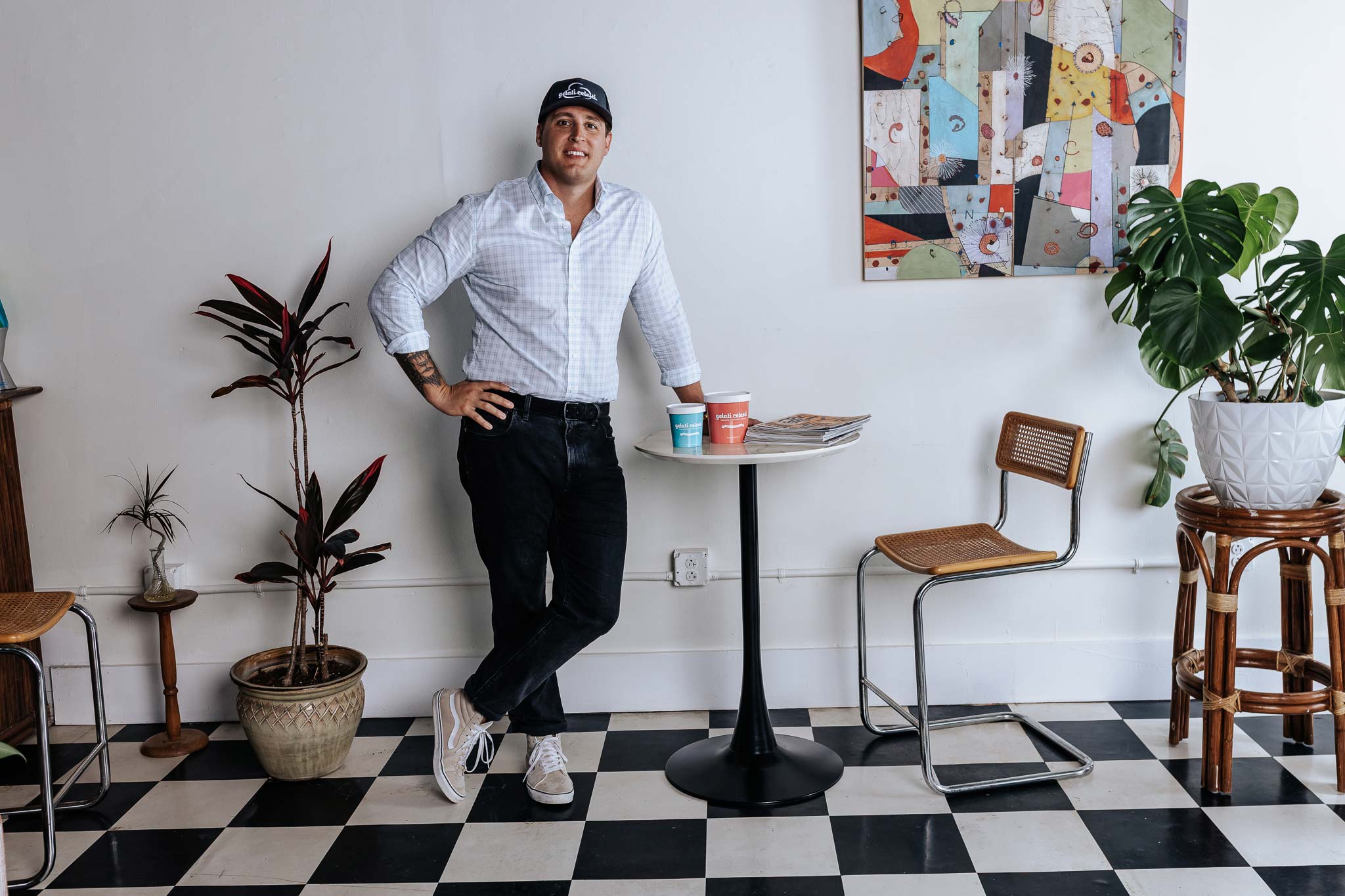Man standing in a cafe with a checkered floor, plants, and a colorful painting on the wall.