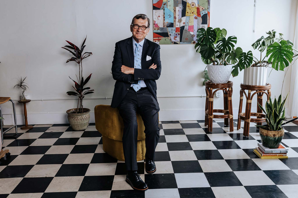 Man in a suit sitting on a chair in a room with checkered floor and plants.