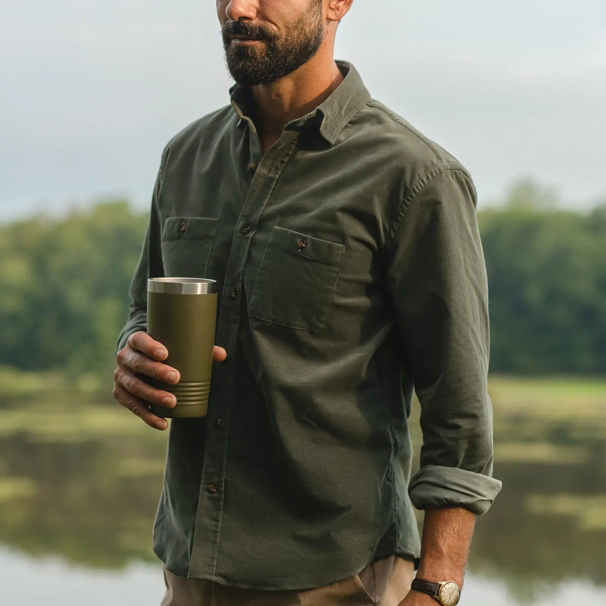 A man outdoors by a lake wearing an olive green long-sleeve button-up shirt and holding an olive green insulated tumbler.