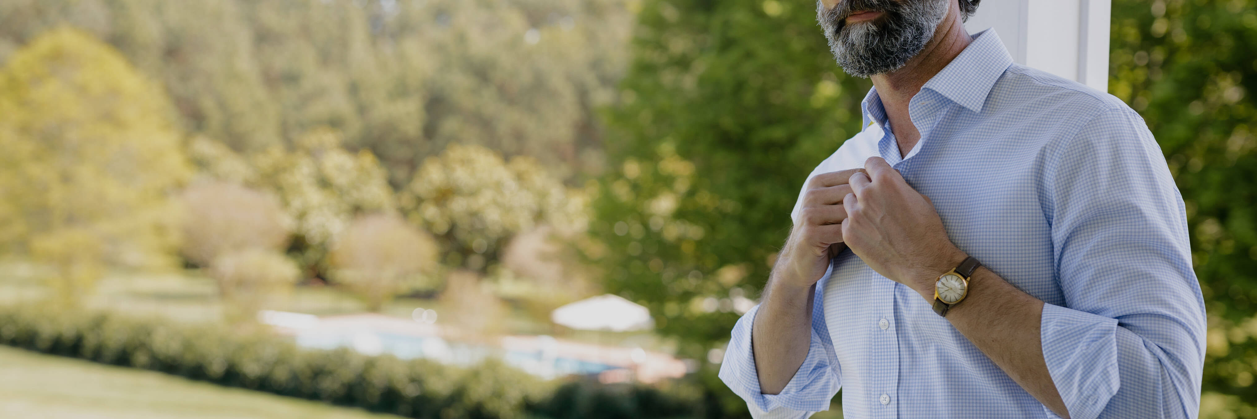 Image of a male model standing on a deck buttoning the neck of a blue gingham button down dress shirt