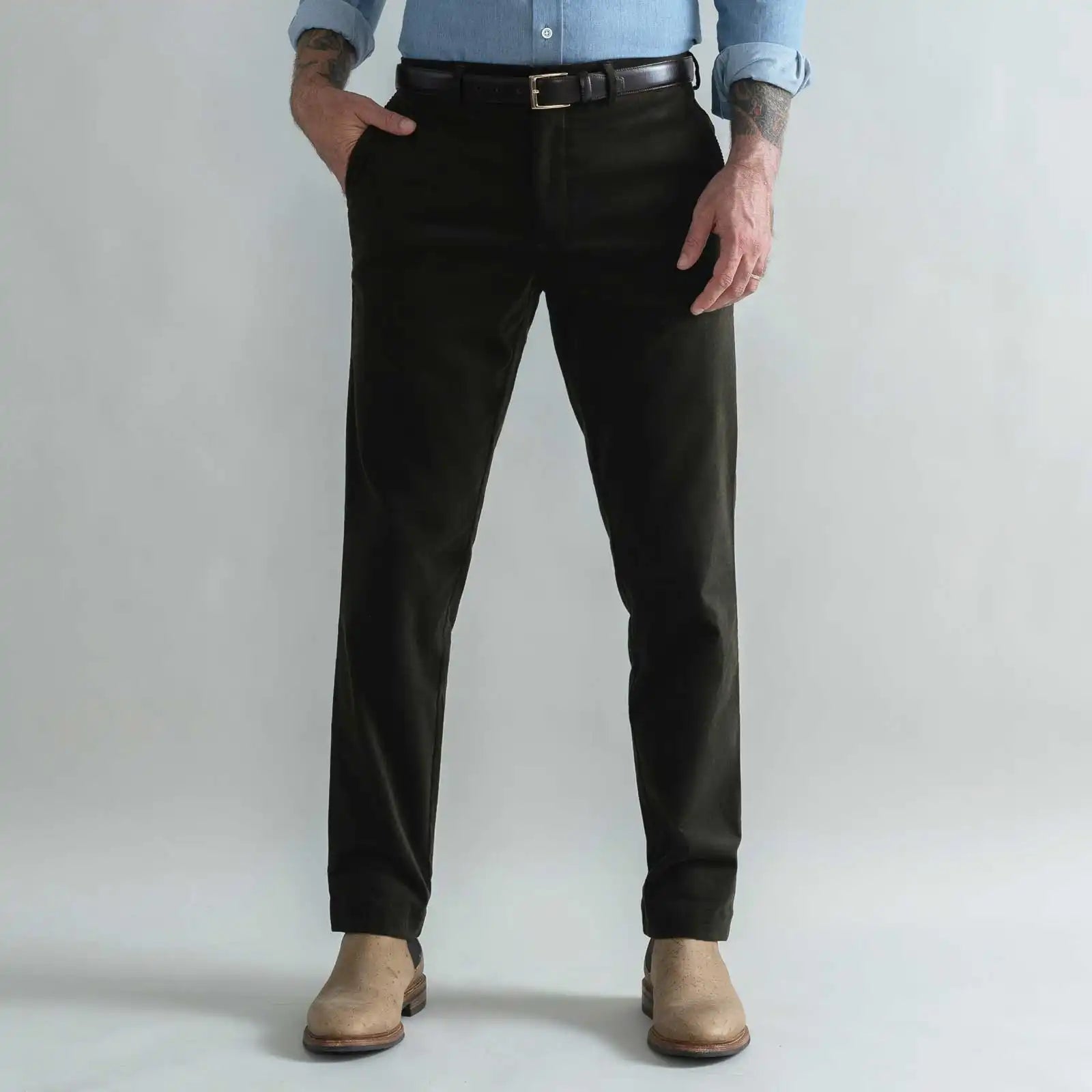 A man wearing slim-fit dark green corduroy trousers, a black belt, a light blue button-down shirt, and tan boots, standing against a light gray studio background.
