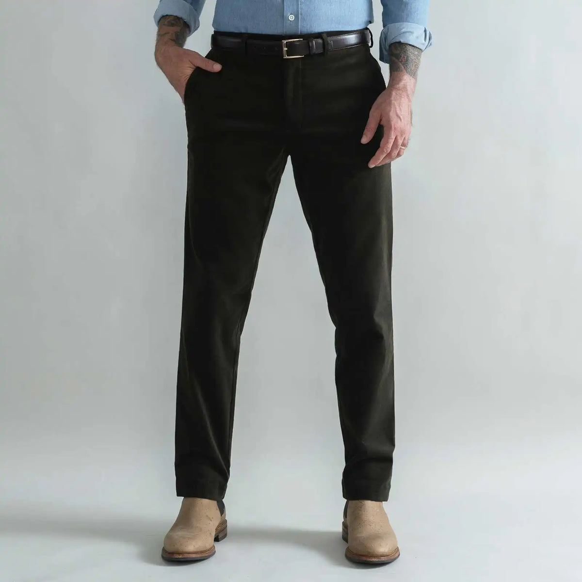 A man wearing slim-fit dark green corduroy trousers, a black belt, a light blue button-down shirt, and tan boots, standing against a light gray studio background.