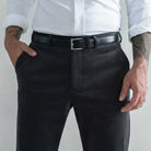 A close-up of a man wearing dark charcoal gray corduroy trousers, a black leather belt with a silver buckle, a white button-down shirt, and tattoos on both arms, set against a plain light gray background.
