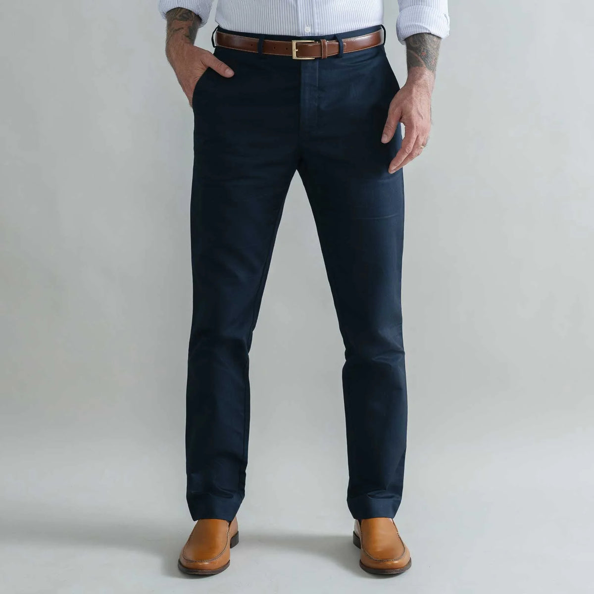 A man wearing dark navy blue trousers, a brown leather belt, a white button-down shirt, and brown leather shoes, standing against a light gray studio background.