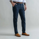 A man wearing slim-fit navy blue trousers, styled with a brown leather belt, a white shirt, and tan leather shoes, standing against a light gray studio background.